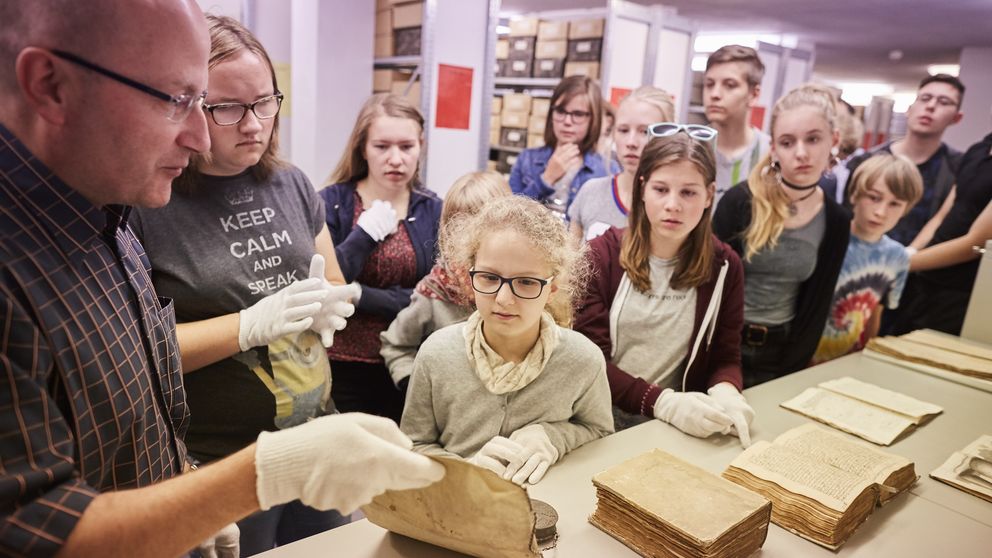 Eine Archivmitarbeiterin zeigt Kindern ein altes Buch und ein dazugehöriges Dokument und erklärt ihnen die historische Bedeutung