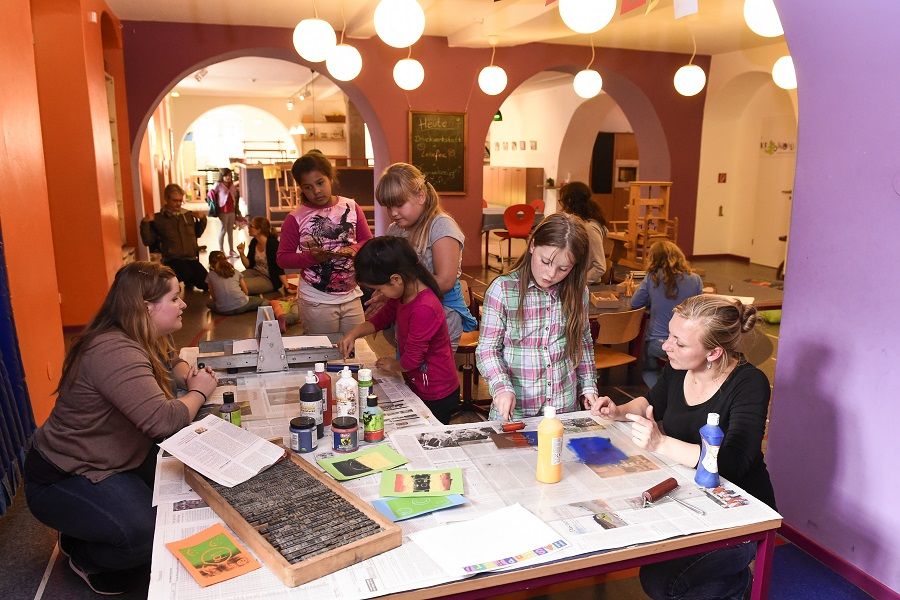 Kinder arbeiten mit Farbe und Druckplatten in der Druckwerkstatt; um sie herum liegen Farben, Schwämme und Setzkästen – kreatives Drucken im Krokoseum