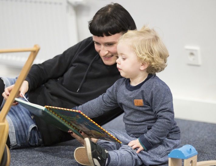 A young woman lies on her side on a carpet, reading a children’s book to a little girl. Together they look at the pictures – a moment of language development and bonding.
