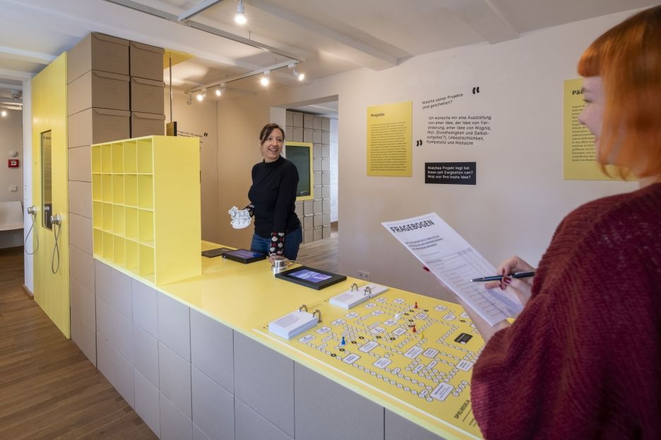 Zwei Besucherinnen erkunden Mitmachstationen in der Ausstellung „Francke im Wandel“ – sie nutzen einen digitalen Objektscanner und spielen ein thematisches Brettspiel zur Reflexion über Geschichte und Gegenwart.